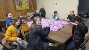 Children sit around a table upon which sits a flowered fleece blanket they are crafting. Students are looking at the camera and smiling.